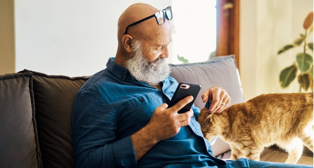 older man with beard looking at social media on his phone on the couch with an orange cat