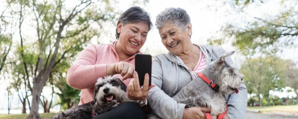 2 older women smiling with their dogs and looking at a mobile phone