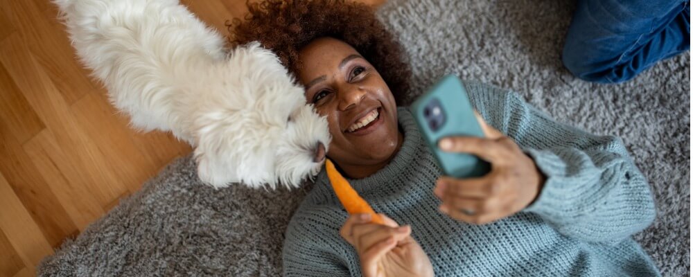 black woman lying on ground smiling and holding her cell phone while a small white dog tries to eat the carrot in her other hand