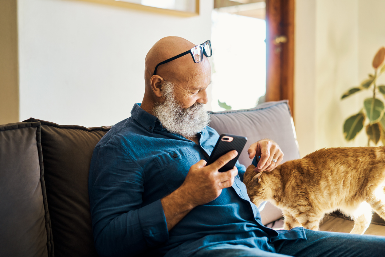 Older man with beard using a mobile device and playing with his orange cat on the couch at home. 