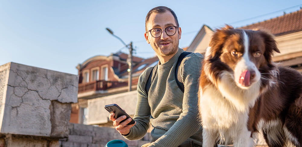 Man sitting on bench outside with mobile phone in hand, looking at his dog standing next to him